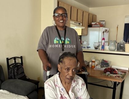 Two people are in an apartment living room and kitchen. An older adult wearing a floral pajama set sits in a wheelchair in the foreground with their hands resting in their lap, while another adult stands behind the wheelchair, smiling and holding the handles. The room includes a couch, chair, folding table with medications and household items, and an open kitchen in the background.