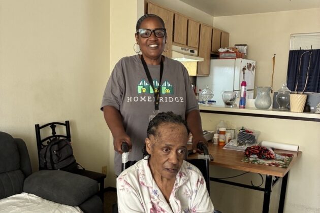 Two people are in an apartment living room and kitchen. An older adult wearing a floral pajama set sits in a wheelchair in the foreground with their hands resting in their lap, while another adult stands behind the wheelchair, smiling and holding the handles. The room includes a couch, chair, folding table with medications and household items, and an open kitchen in the background.