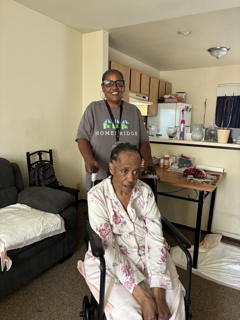 Two people are in an apartment living room and kitchen. An older adult wearing a floral pajama set sits in a wheelchair in the foreground with their hands resting in their lap, while another adult stands behind the wheelchair, smiling and holding the handles. The room includes a couch, chair, folding table with medications and household items, and an open kitchen in the background.